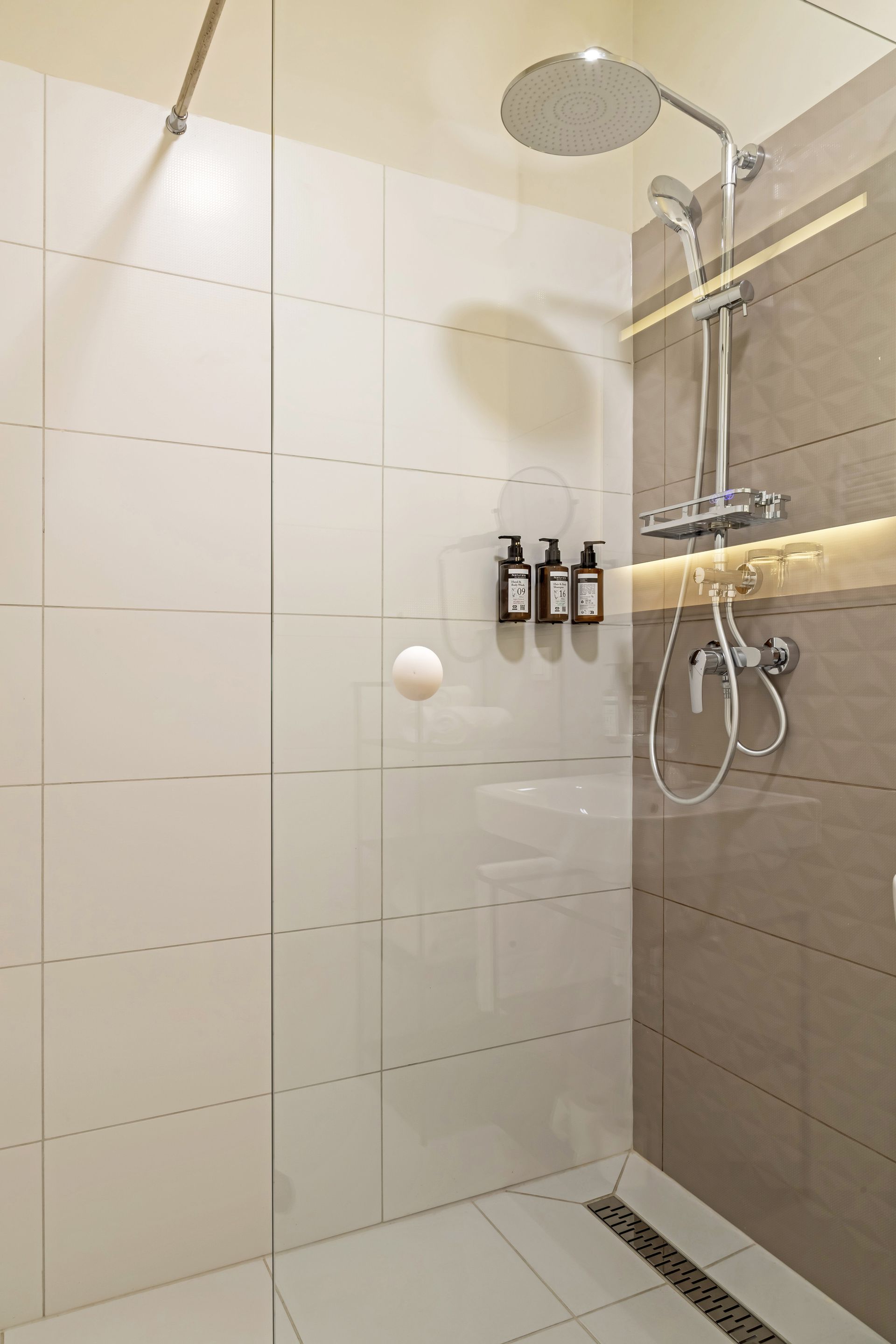 Walk-in shower with white and gray tiled walls, rain shower head, shower gel, shampoo and conditioner bottles. Marone Suites