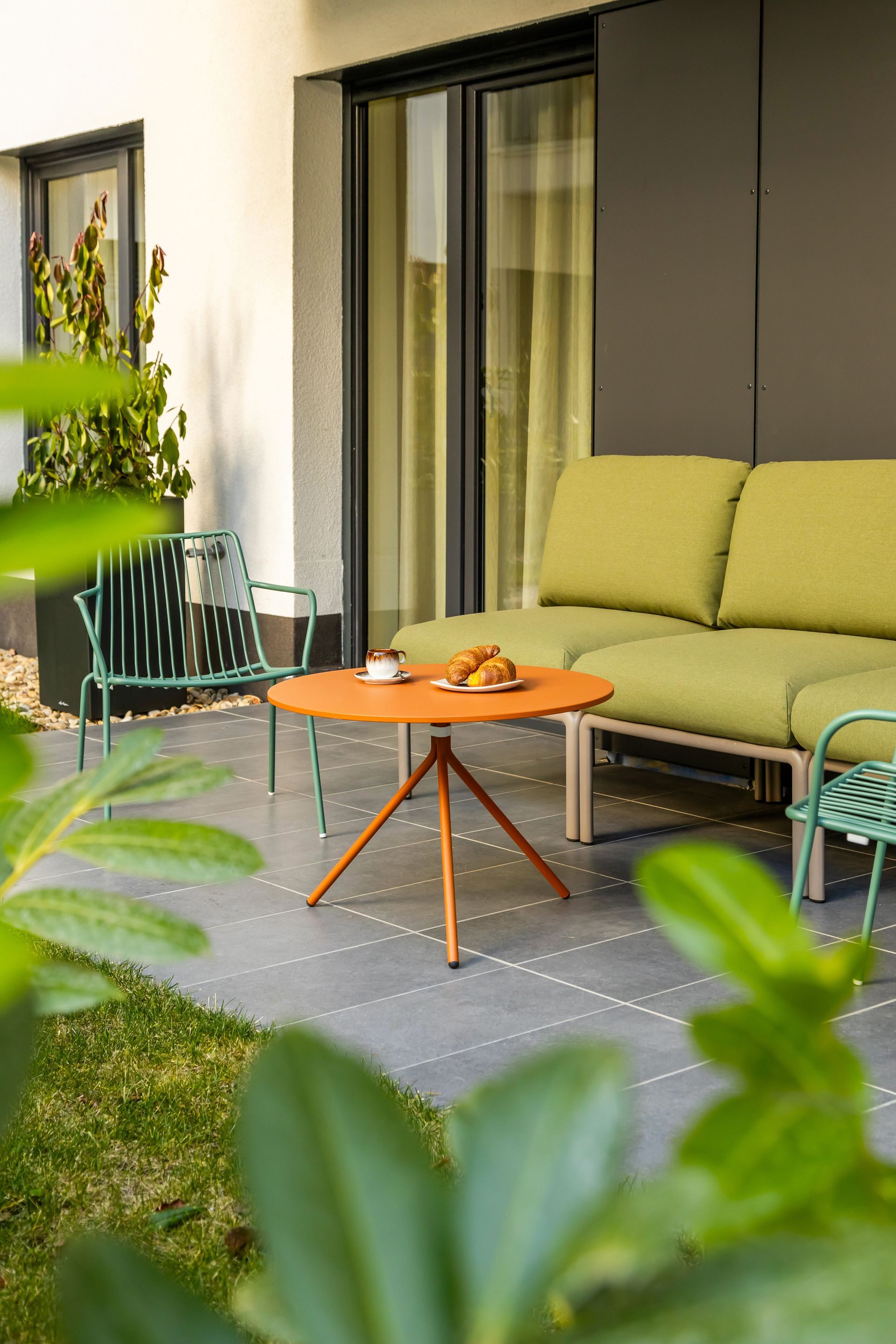 Outdoor seating area with orange table, green sofa and chairs, and a glass door in Marone Suites Budapest.
