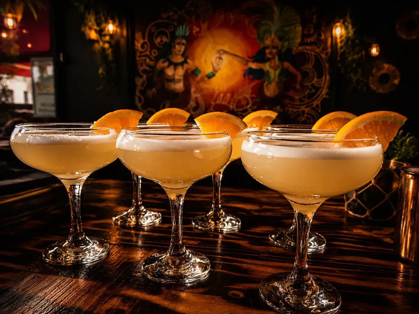 Tres cócteles coupe adornados con rodajas de naranja en una barra pulida en un salón con luz tenue.
