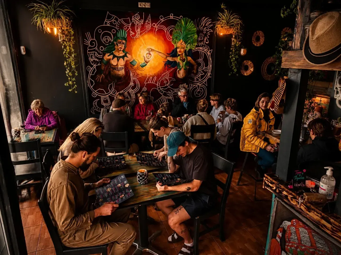 Cafetería tropical abarrotada de gente comiendo bajo murales de color naranja cálido y plantas colgantes.