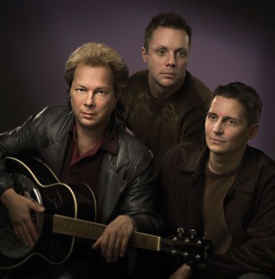 Kolme miestä poseeraa kuvaa varten ja yksi heistä pitelee kitaraa