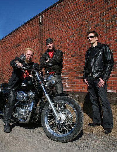 Kolme miestä seisoo moottoripyörän vieressä tiiliseinän edessä