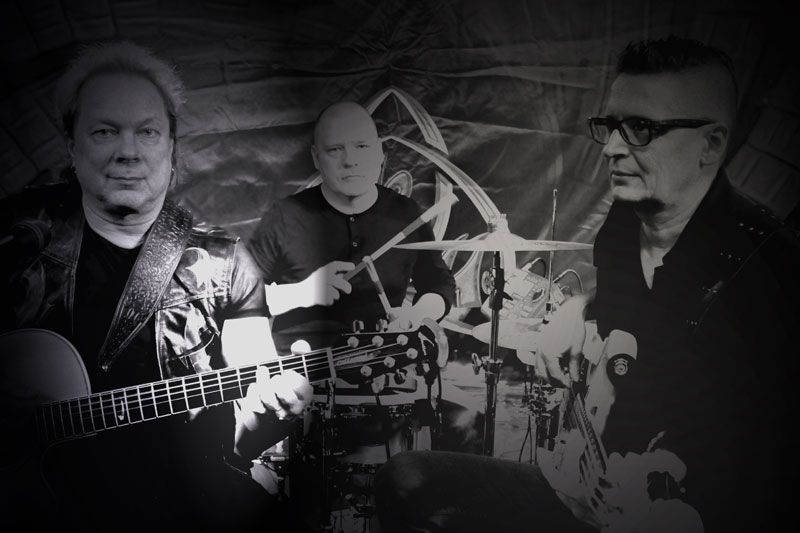 Kolme miestä soittaa kitaroita ja rumpuja mustavalkoisessa valokuvassa.