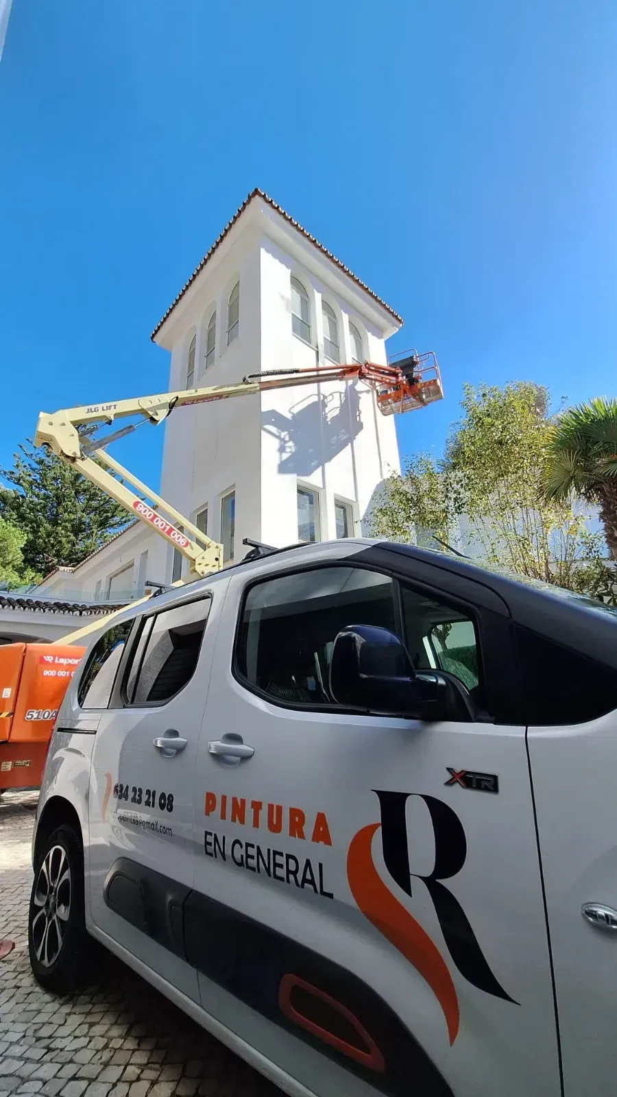 Un edificio blanco siendo pintado por un trabajador en un elevador aéreo, con una camioneta blanca en primer plano.