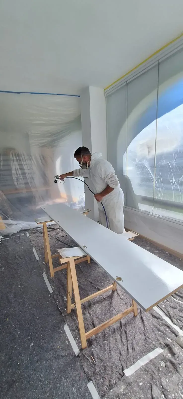 Una persona con un traje protector rocía pintura sobre una tabla larga y blanca sostenida por caballetes en una habitación.