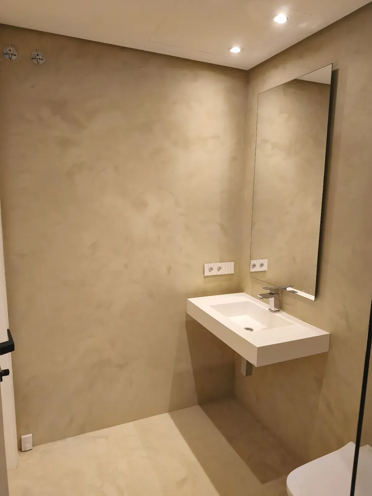 Baño moderno con paredes texturizadas en color beige, lavabo flotante, espejo grande e iluminación empotrada.