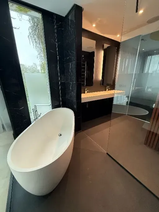 Baño moderno con bañera independiente blanca, pared de mármol negro, ducha de cristal y tocador doble.