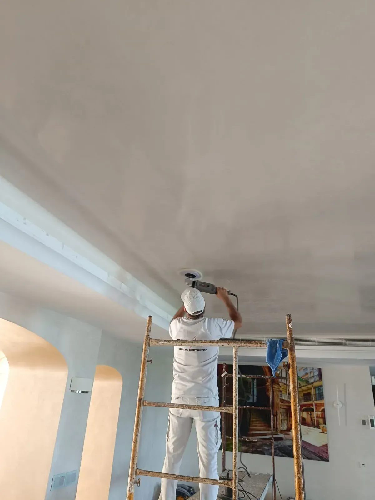Persona en una escalera lijando un techo, vestida con un mono blanco, en un espacio interior.