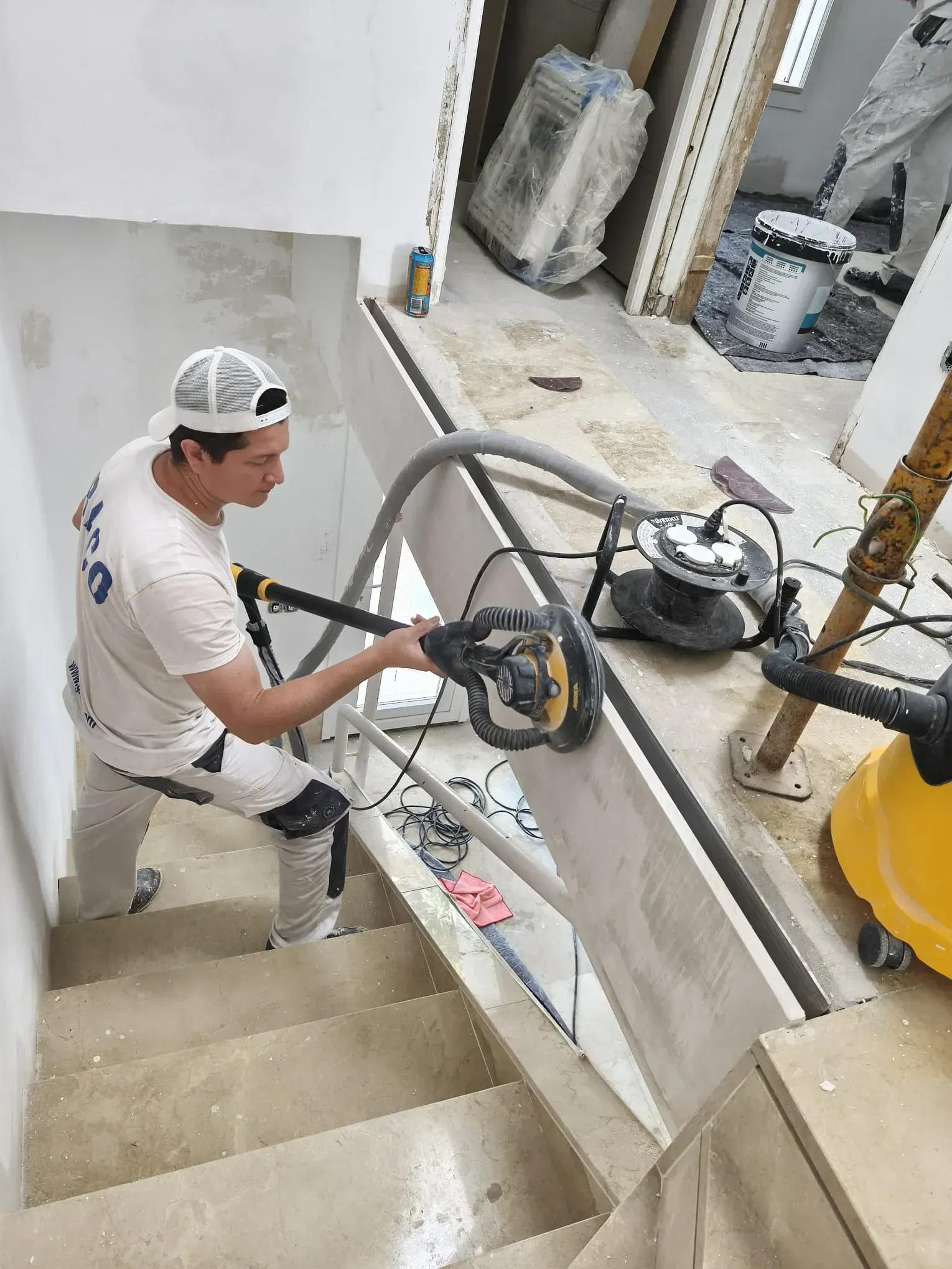 Hombre lijando una barandilla de escalera, vestido con gorra y mono, con una lijadora conectada a una aspiradora.