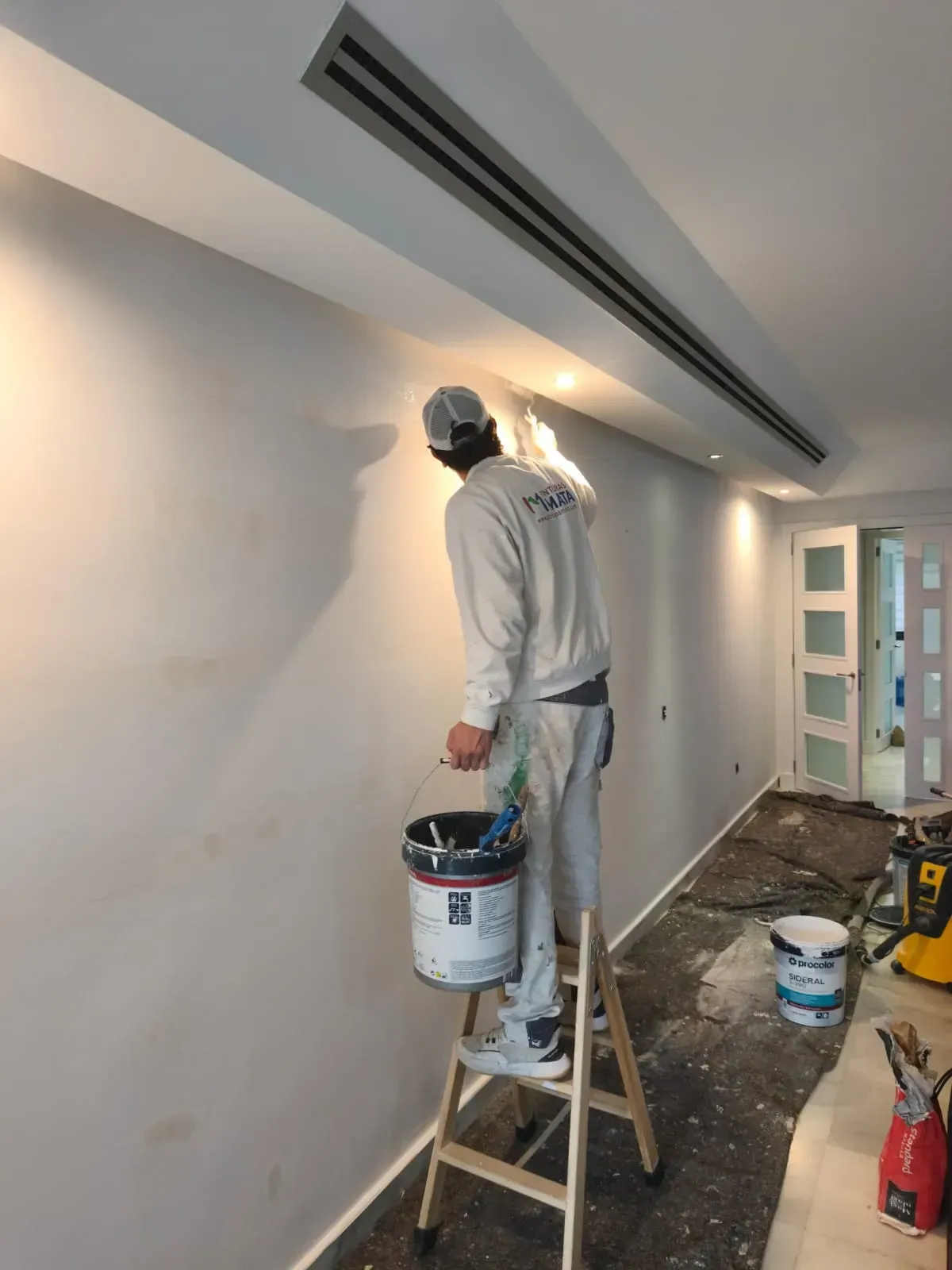 Pintor en una escalera, aplicando pintura a una pared blanca en una habitación con un respiradero largo.