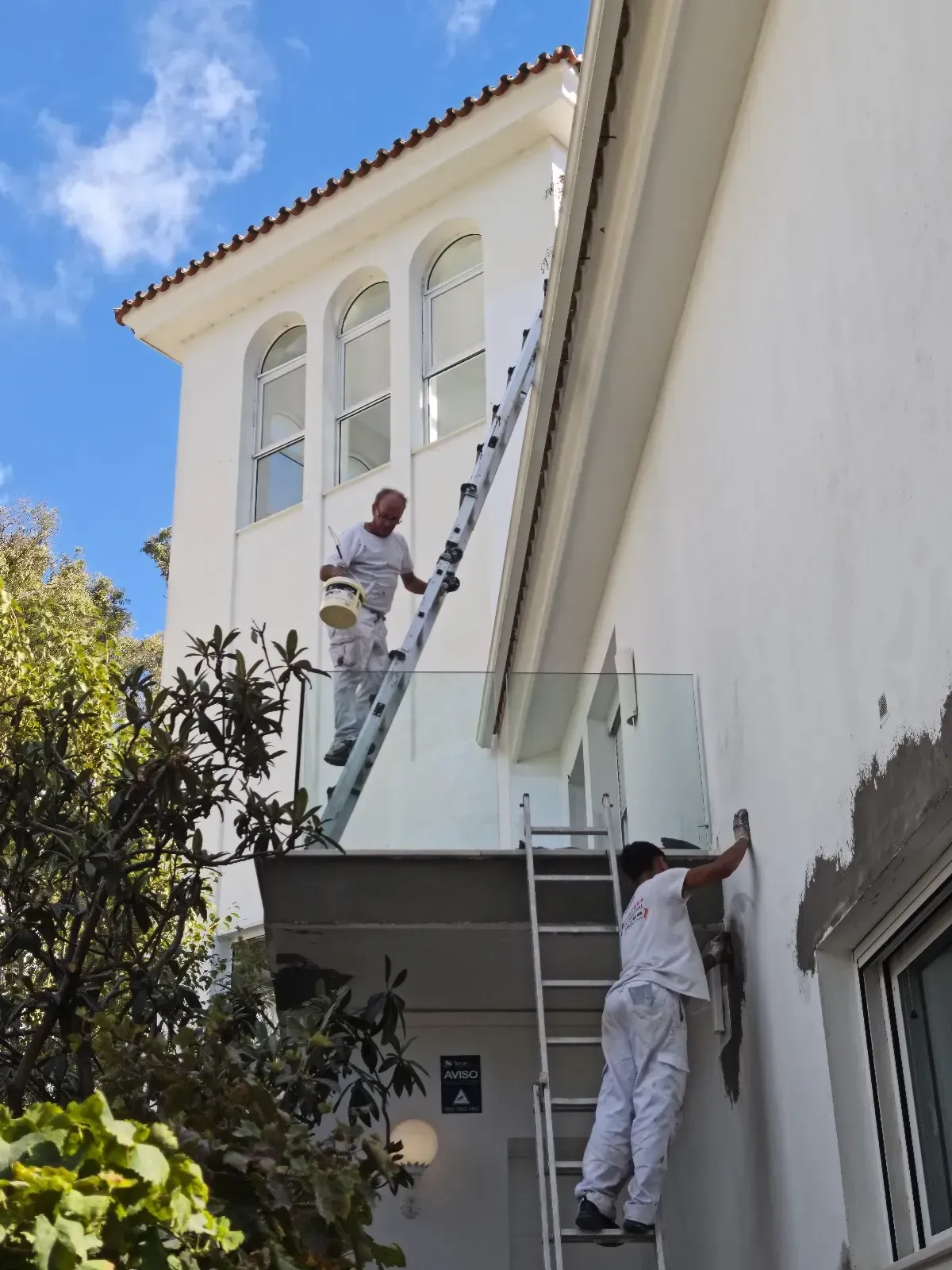 Dos pintores con overoles blancos pintan un edificio blanco en un día soleado. Uno en una escalera, el otro en el suelo.