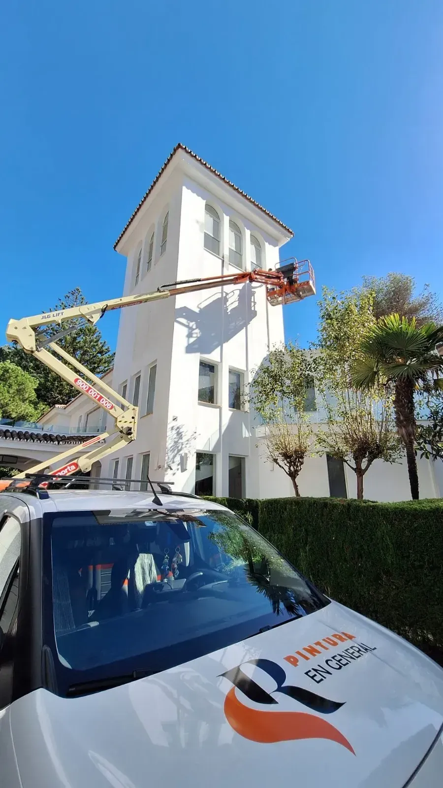 Edificio blanco siendo pintado por un trabajador en un ascensor, día soleado.