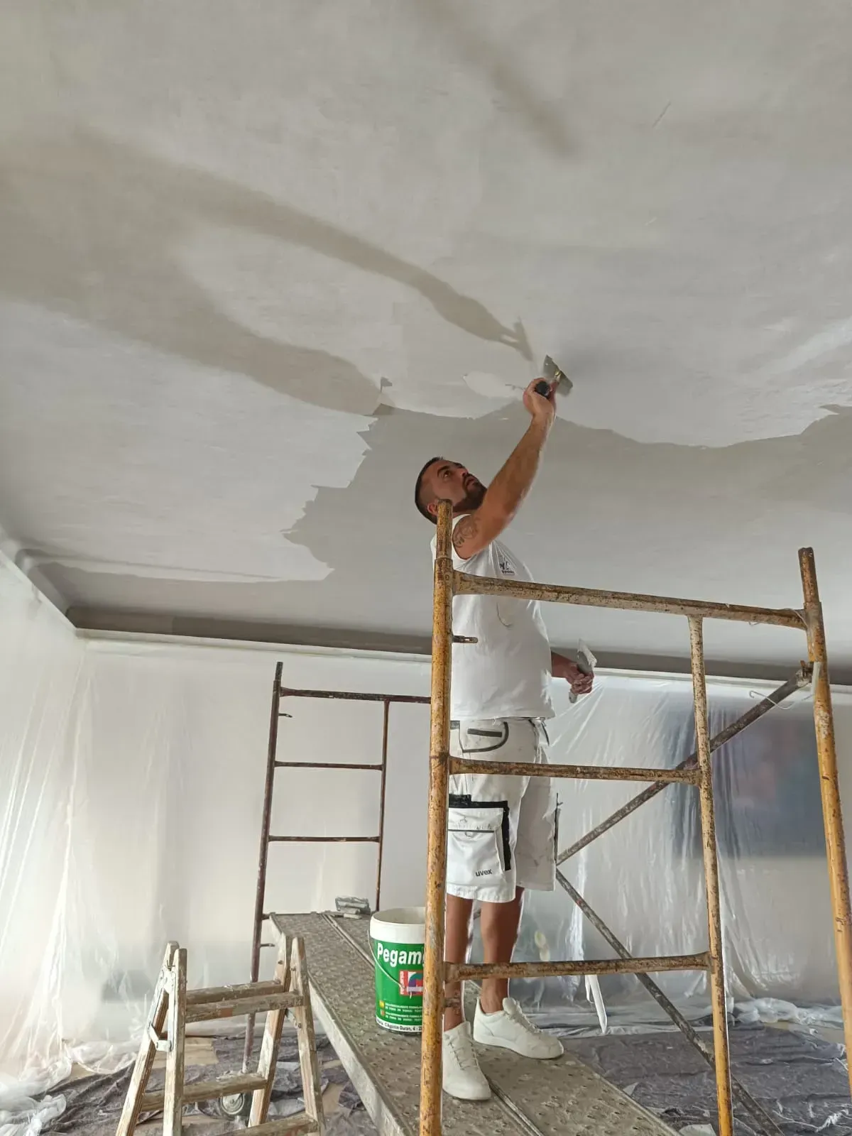 Una persona enyesa un techo mientras está de pie sobre un andamio, con un cubo de pintura cerca, en el interior.