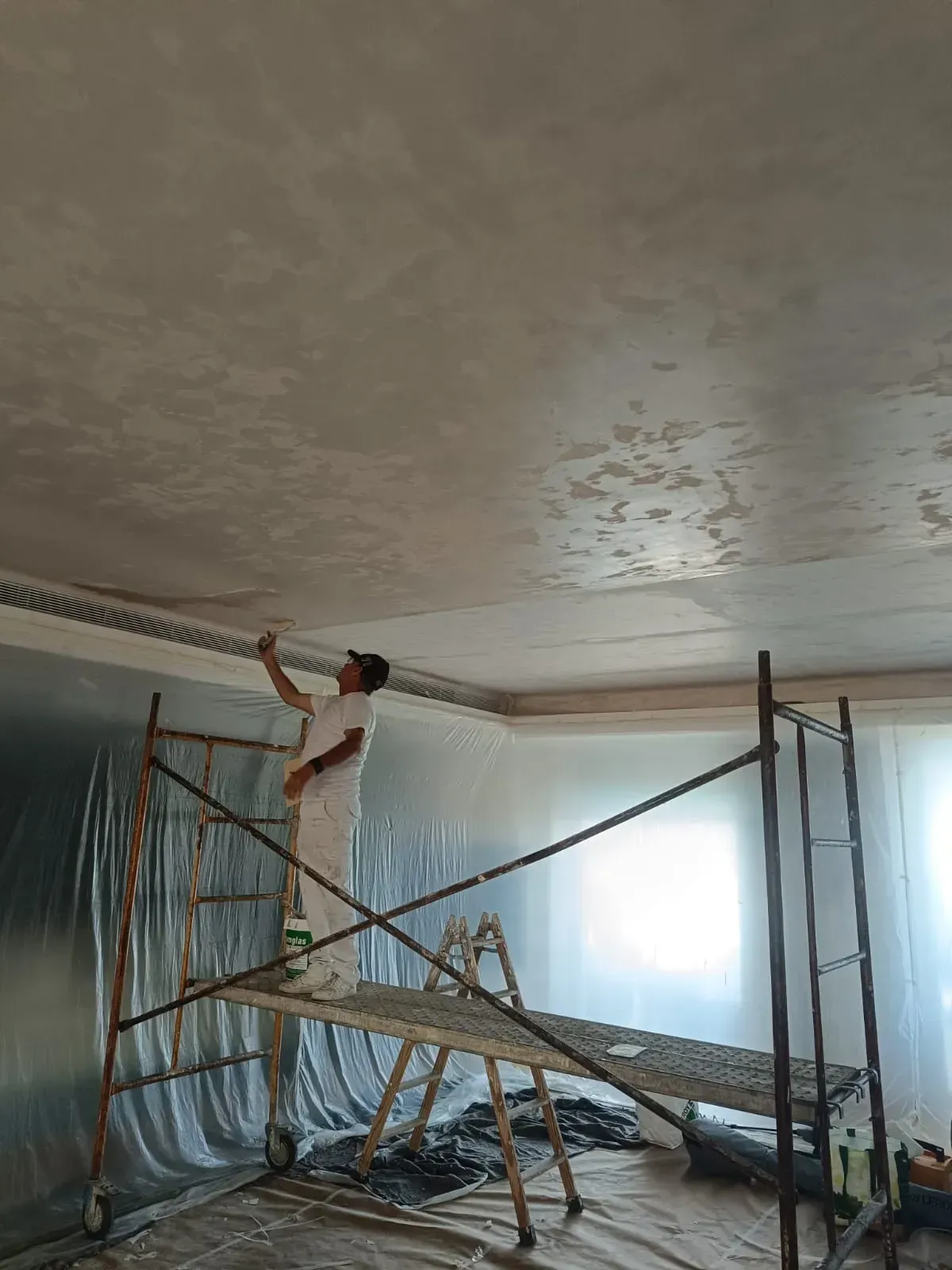 Persona en un andamio pintando un techo en el interior, probablemente aplicando un acabado de yeso.