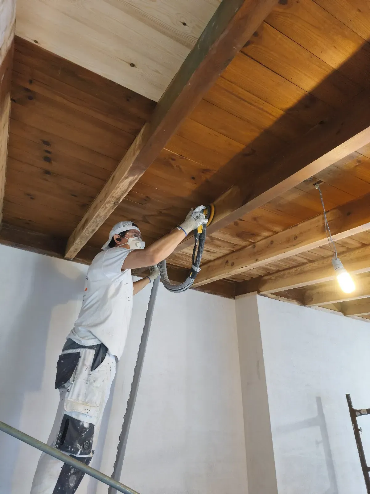Persona lijando vigas de madera del techo, con mascarilla y ropa protectora. En el interior, trabajando desde una escalera.