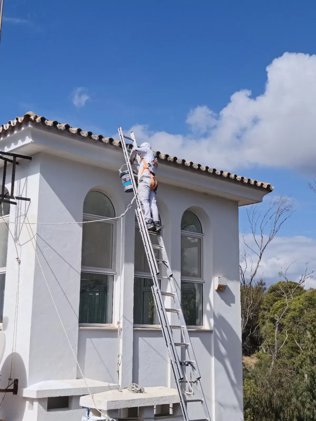 Un pintor subido a una escalera pinta la fachada blanca de un edificio. Día soleado con cielo azul y algunas nubes.
