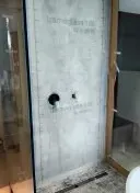 Cabina de ducha con panel posterior impermeable gris, desagüe y puerta de cristal. Fontanería visible.