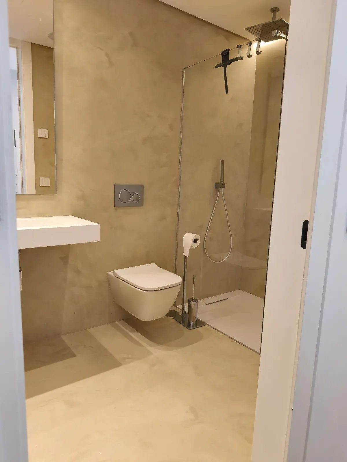 Baño moderno con inodoro suspendido, ducha con mampara de cristal y lavabo blanco. Paredes y suelo beige.