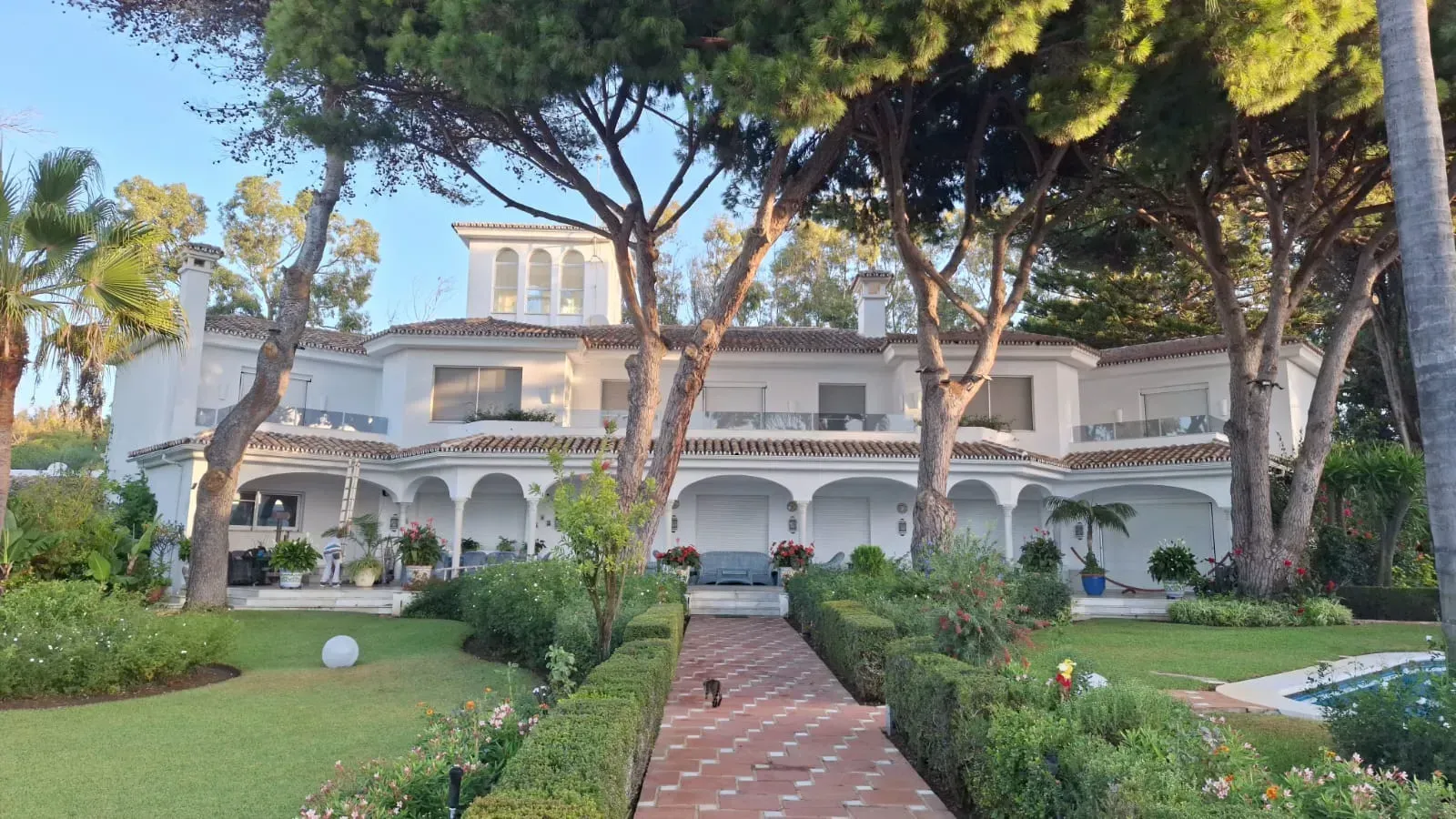 Mansión blanca con pasarela de baldosas rojas, rodeada de exuberante césped verde y árboles.