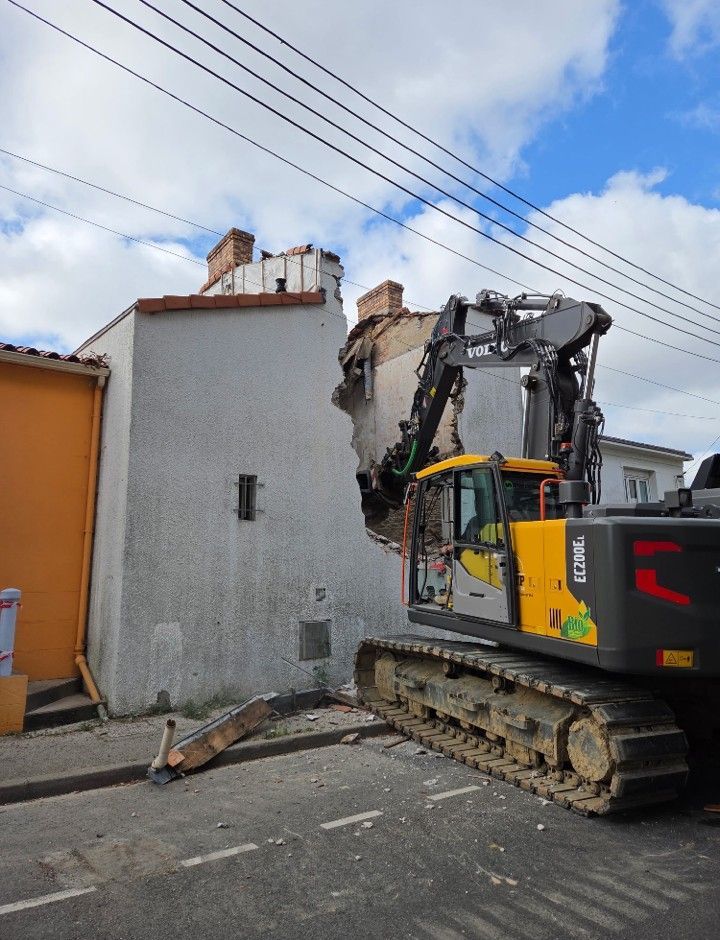 Une pelleteuse démolit un bâtiment blanc dans une rue sous un ciel bleu et des lignes électriques.