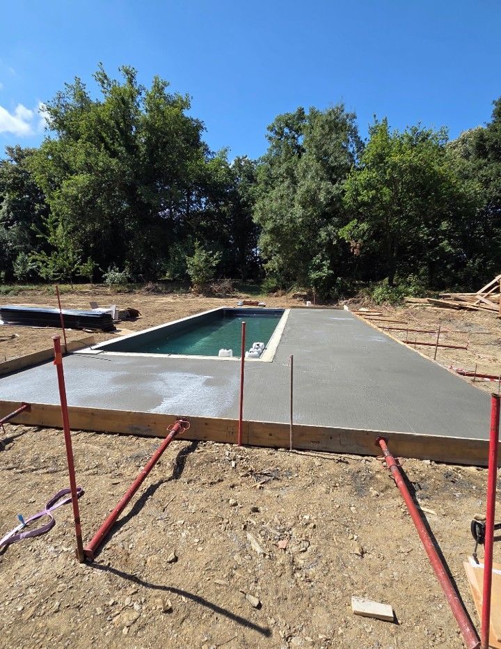Terrasse de piscine en béton en construction à côté d'une piscine de jardin, avec des arbres et un ciel bleu dégagé.