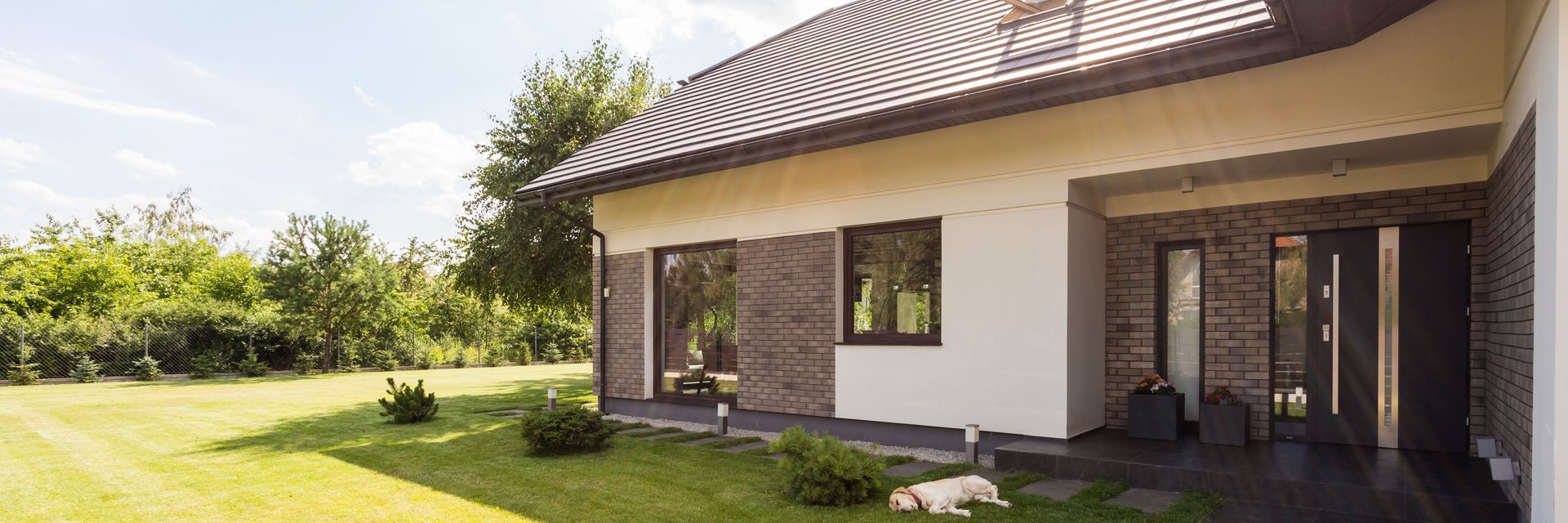 Maison moderne avec façade en pierre, toit brun et grandes fenêtres donnant sur une pelouse verdoyante.