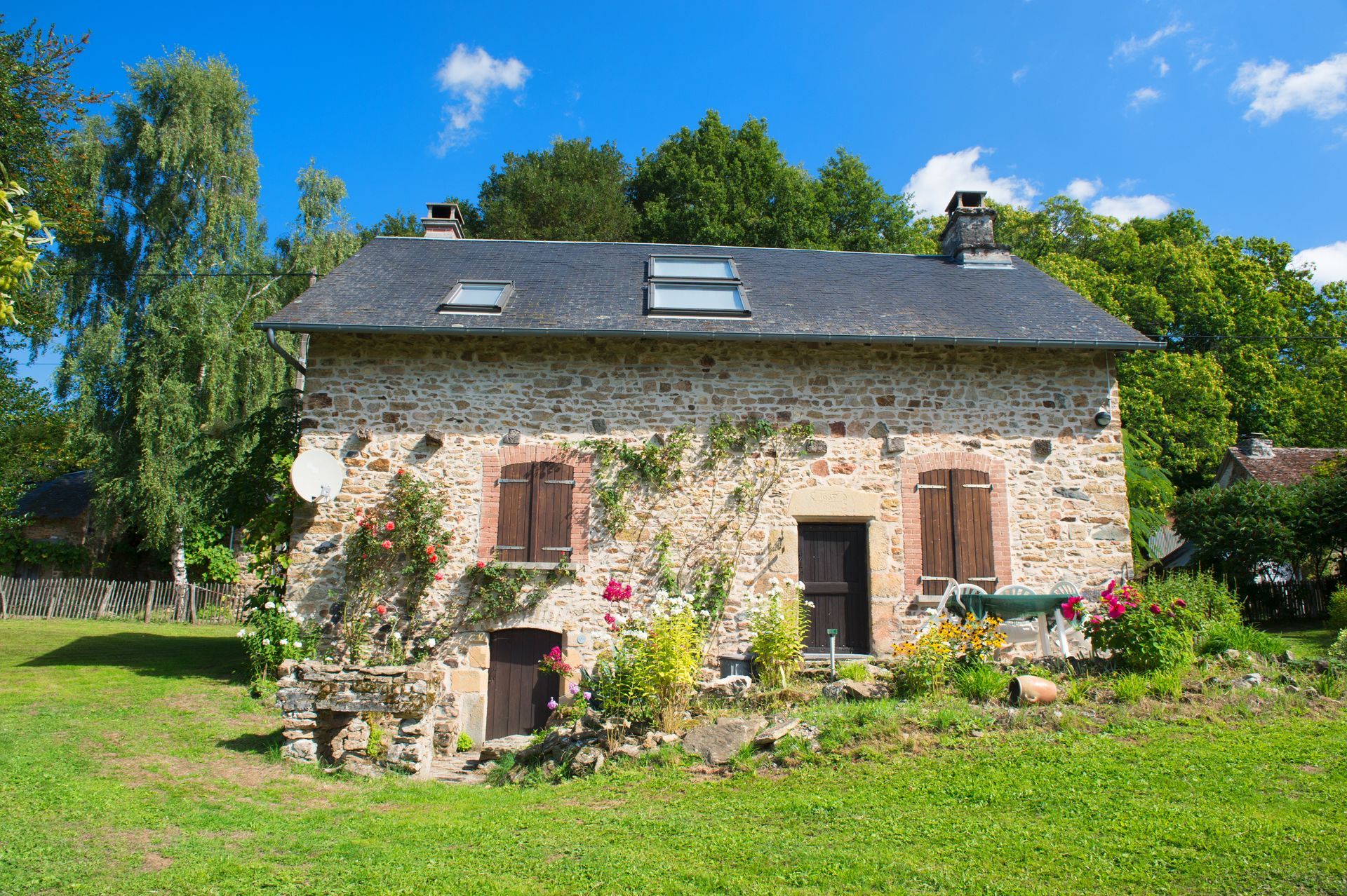 Maison en pierre au toit sombre, jardin fleuri et pelouse verte sous un ciel bleu