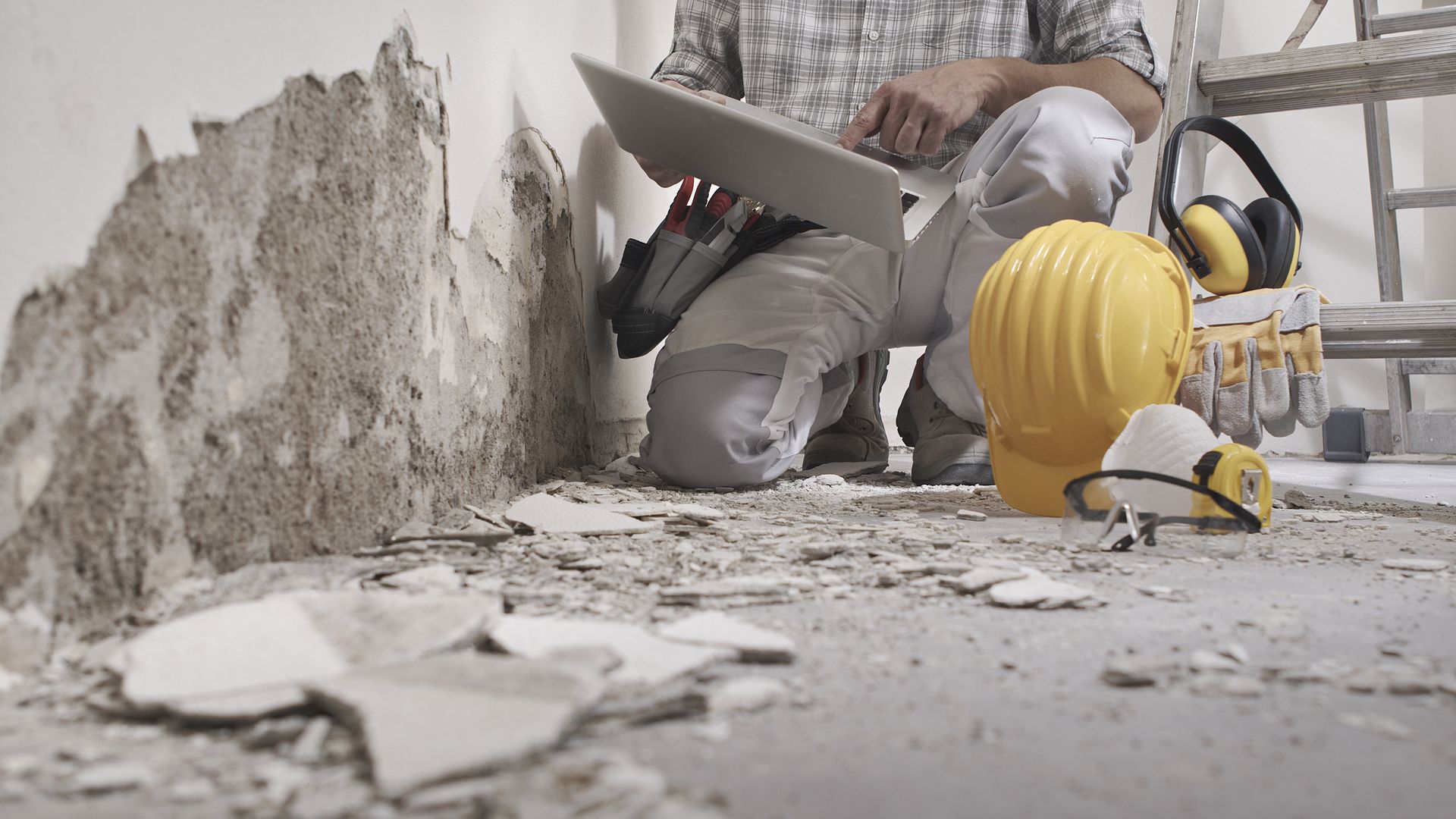 Un ouvrier du bâtiment perce un mur,un casque jaune et ses outils éparpillés sur un sol poussiéreux.