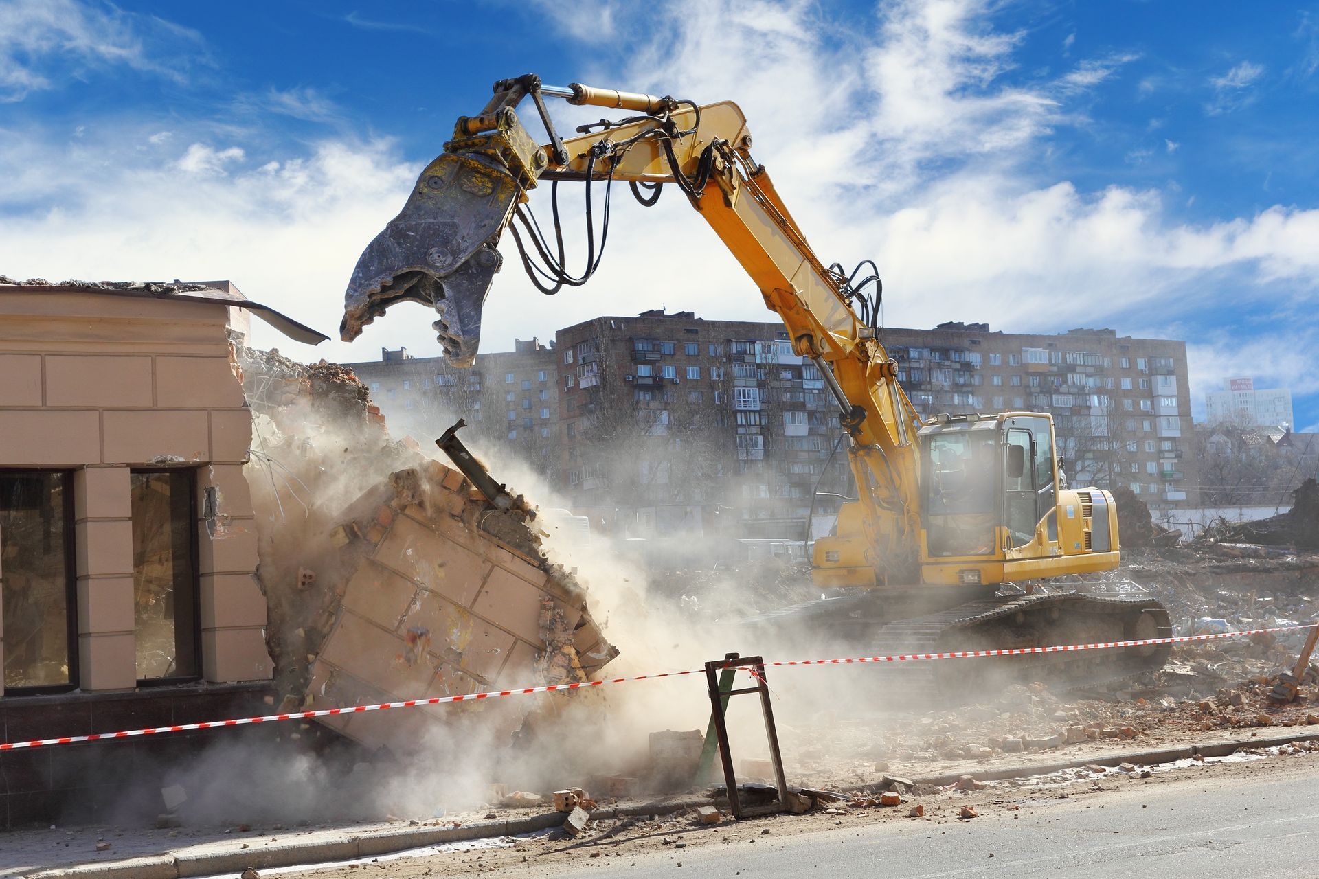 Une pelleteuse démolit un mur de briques, soulevant de la poussière sur un chantier.
