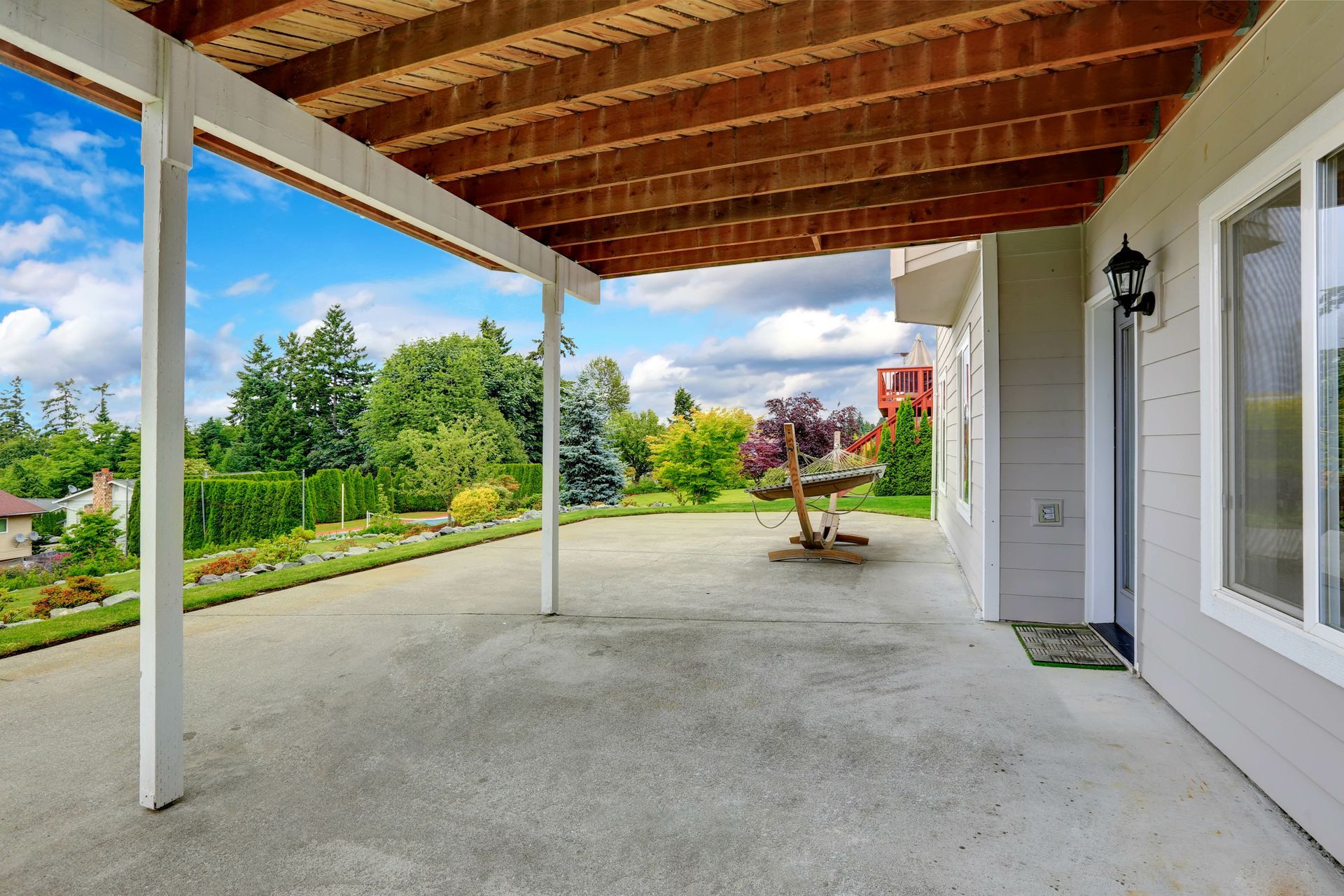 Terrasse couverte en béton avec poteaux blancs, plafond en bois apparent et vue sur le jardin.