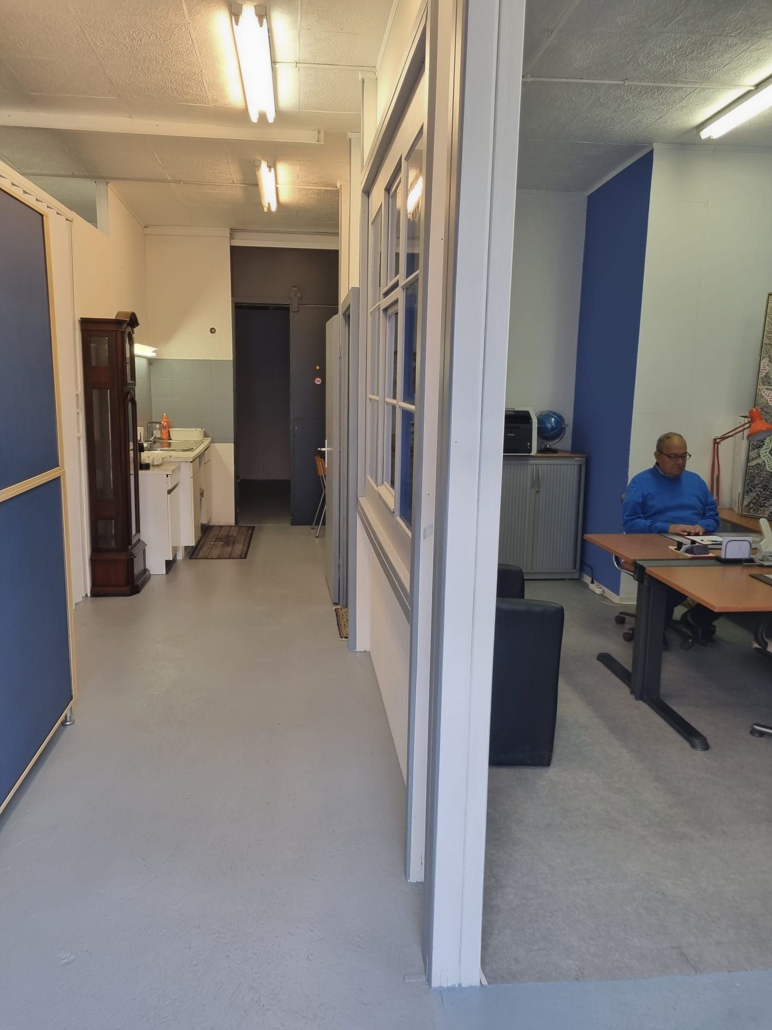 Espace de bureau intérieur, tons gris et bleu. Un homme assis à un bureau. Un couloir mène à une autre pièce.