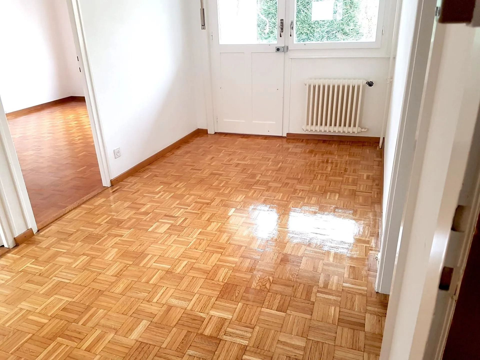 Pièce vide avec parquet, porte blanche, fenêtre et radiateur. Une autre pièce est visible.