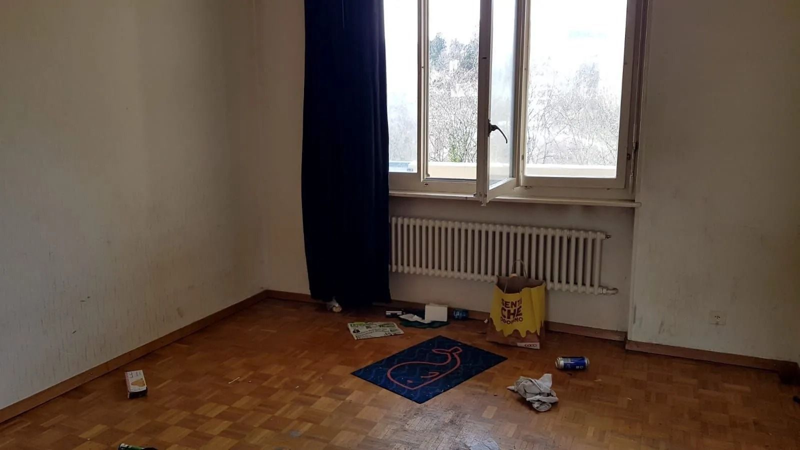 Pièce vide avec parquet sale, fenêtre ouverte, radiateur et déchets éparpillés.