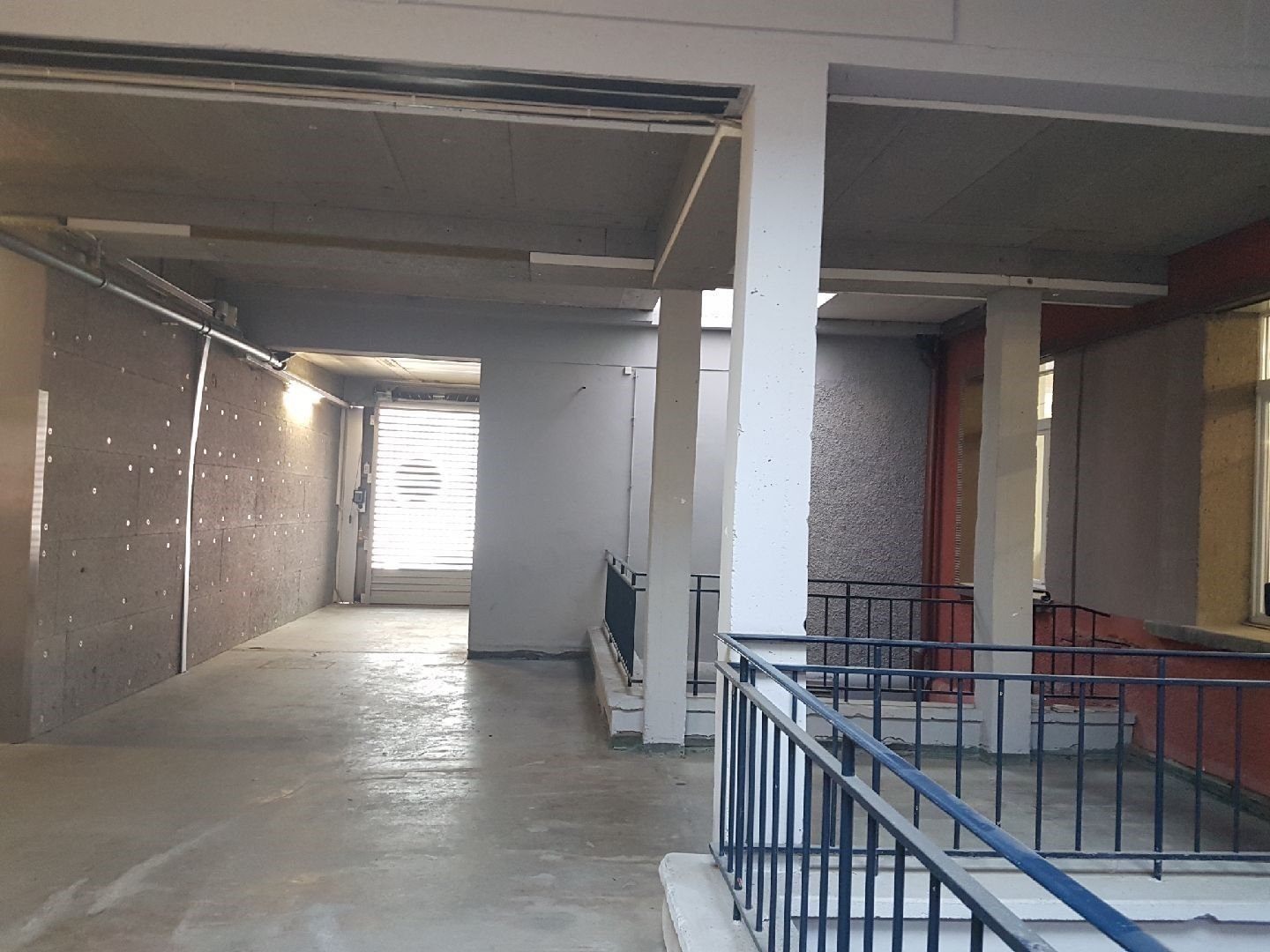 Couloir intérieur avec murs en béton, piliers et balustrade bleue.