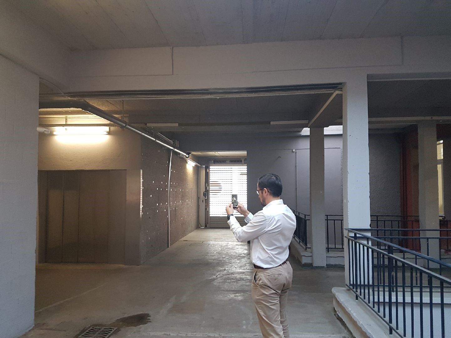 Un homme prend une photo dans un couloir en béton avec un ascenseur, des murs gris et une porte ouverte.