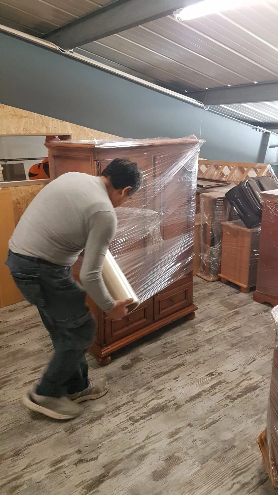 Un homme enveloppe une armoire en bois avec du film plastique dans un entrepôt.