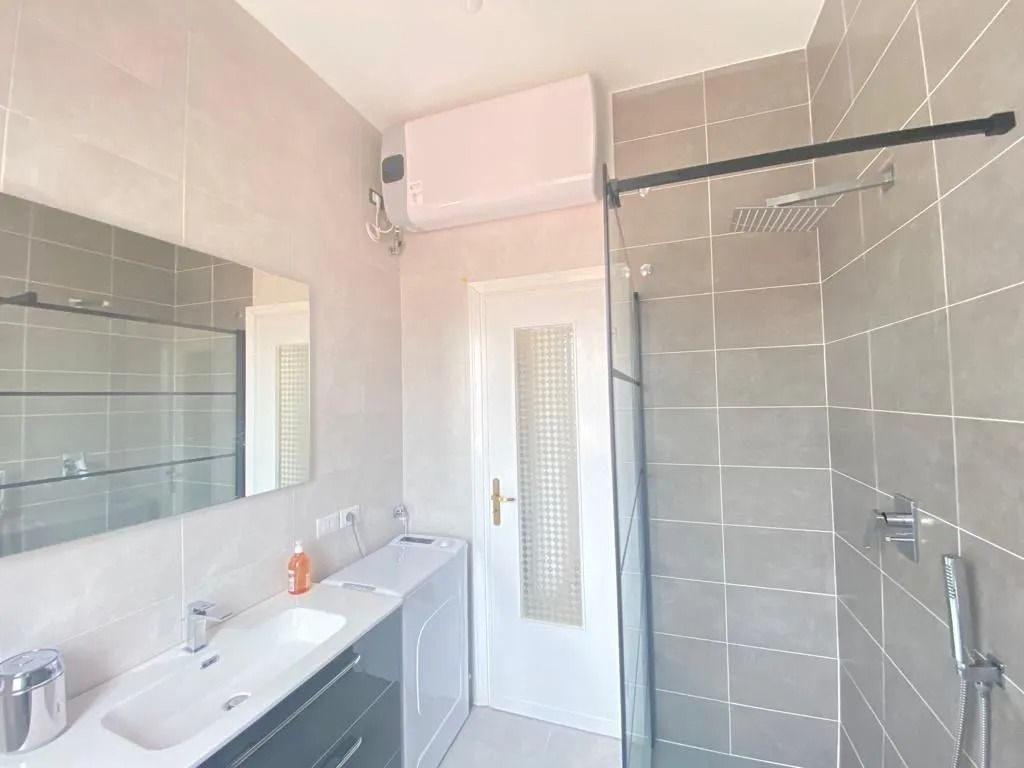 Salle de bains avec carrelage gris, douche, lavabo, miroir et porte blanche. Unité de climatisation au-dessus de la porte.