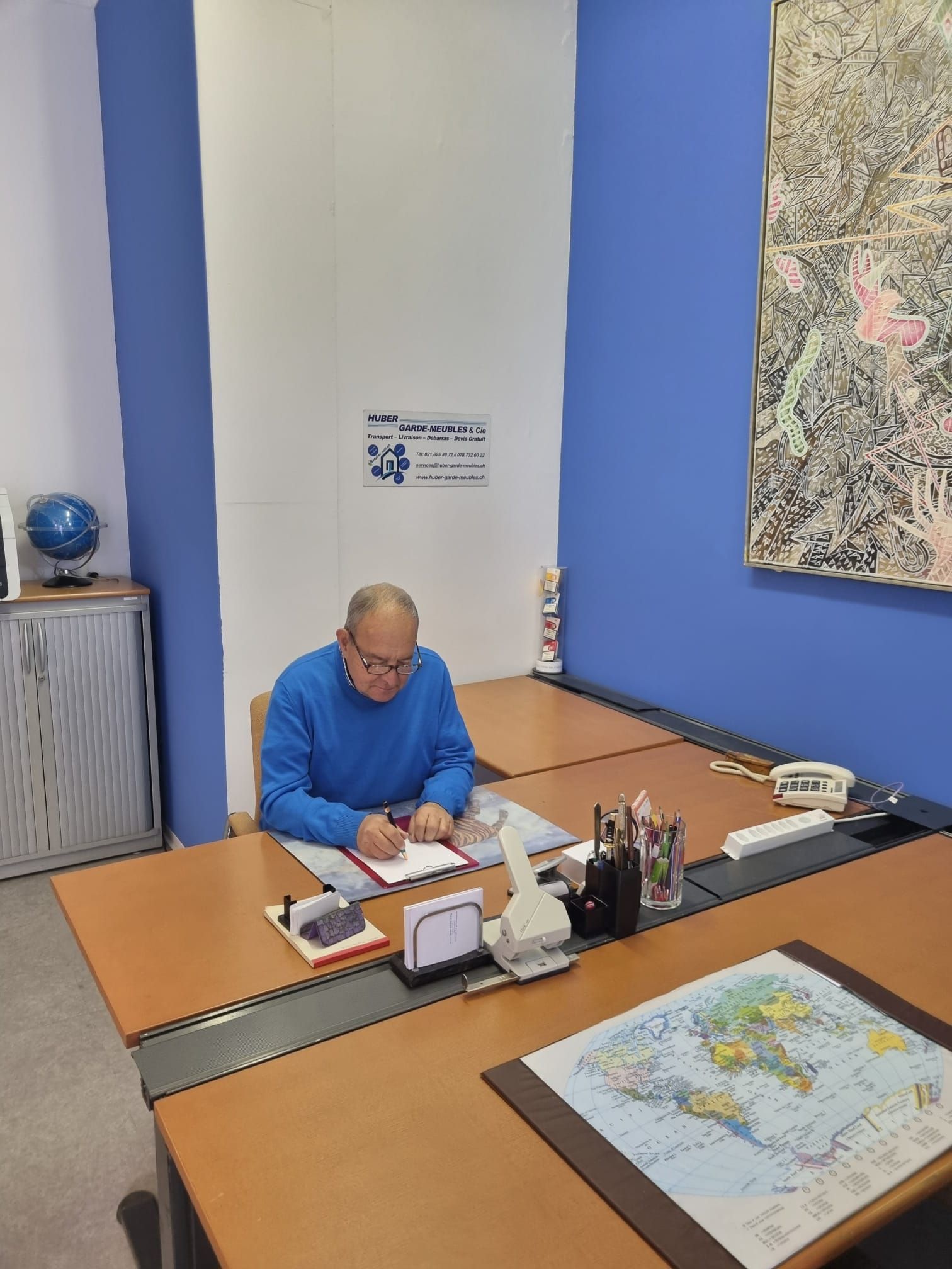 Un homme à son bureau écrit dans un bureau, portant un pull bleu, avec une carte et un globe visibles.