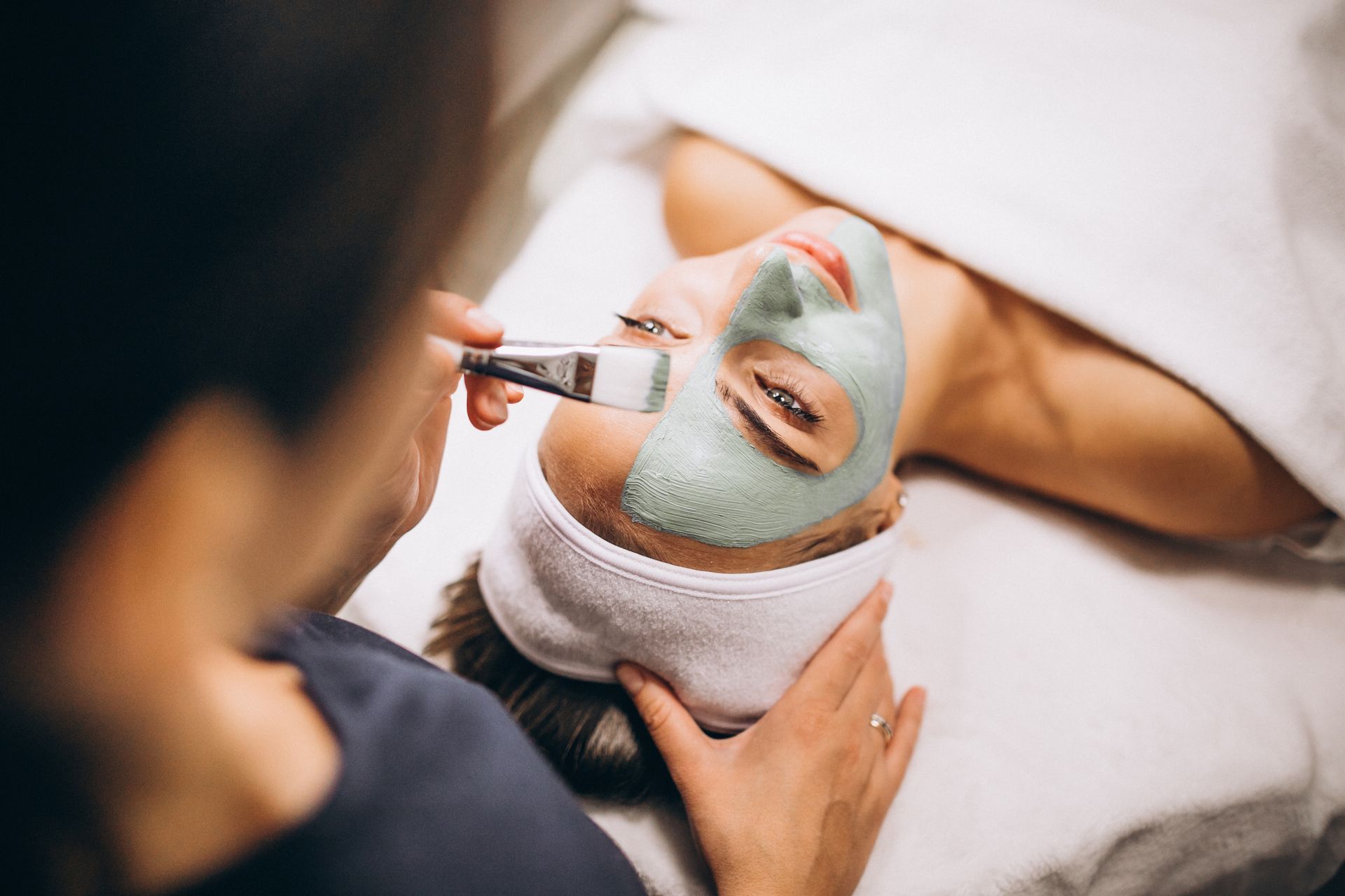 Une personne recevant un masque facial appliqué au pinceau dans un spa, allongée sur le dos avec une serviette enroulée autour de la tête.