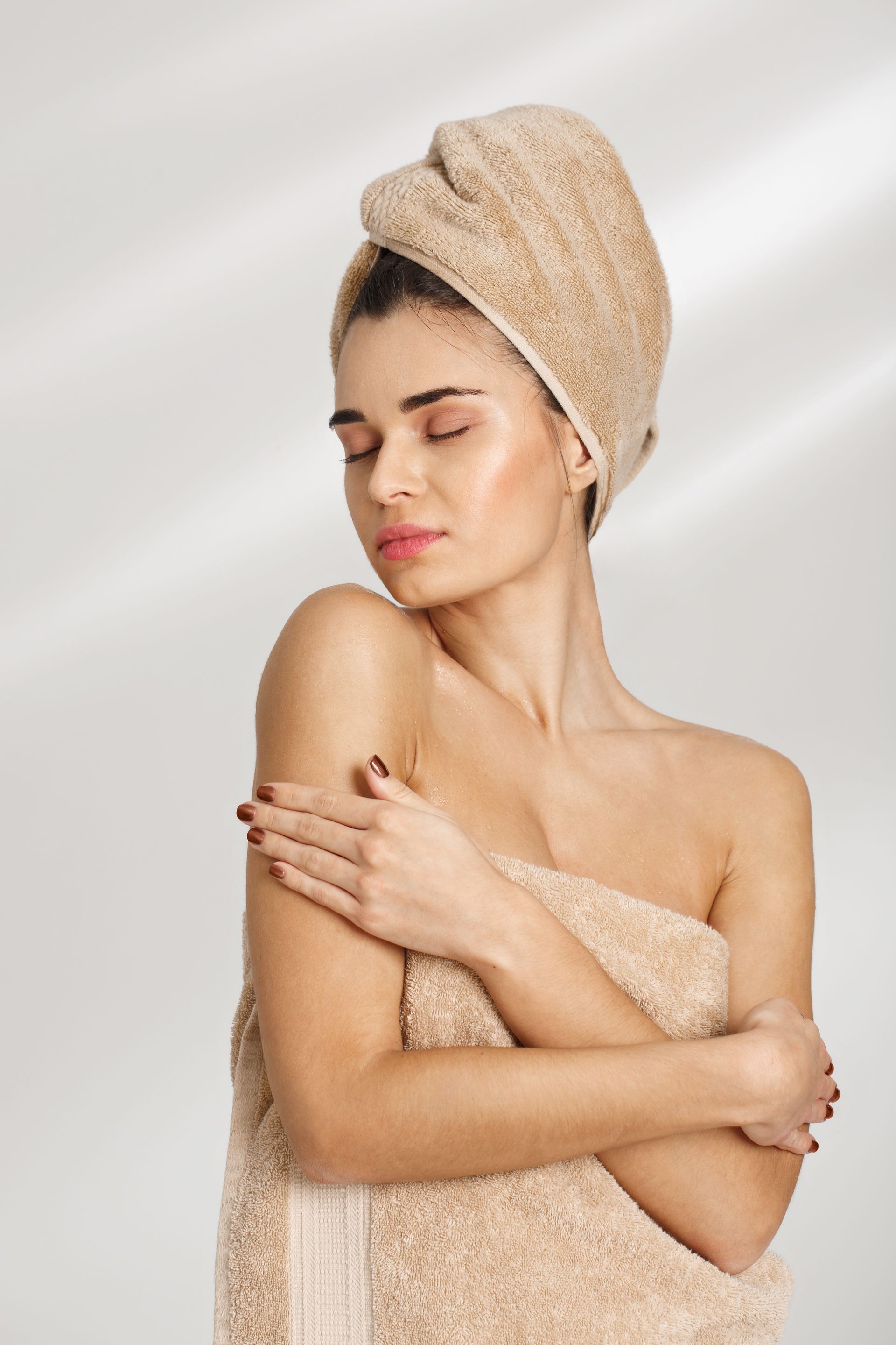Femme enveloppée dans une serviette beige, une serviette sur la tête, se serrant doucement contre elle-même sur un fond blanc doux.