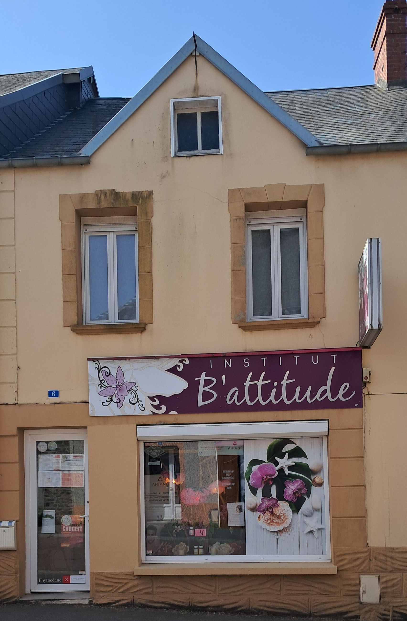 Vue de rue d'une petite boutique beige avec une enseigne rose et un arrangement floral dans une rue tranquille.