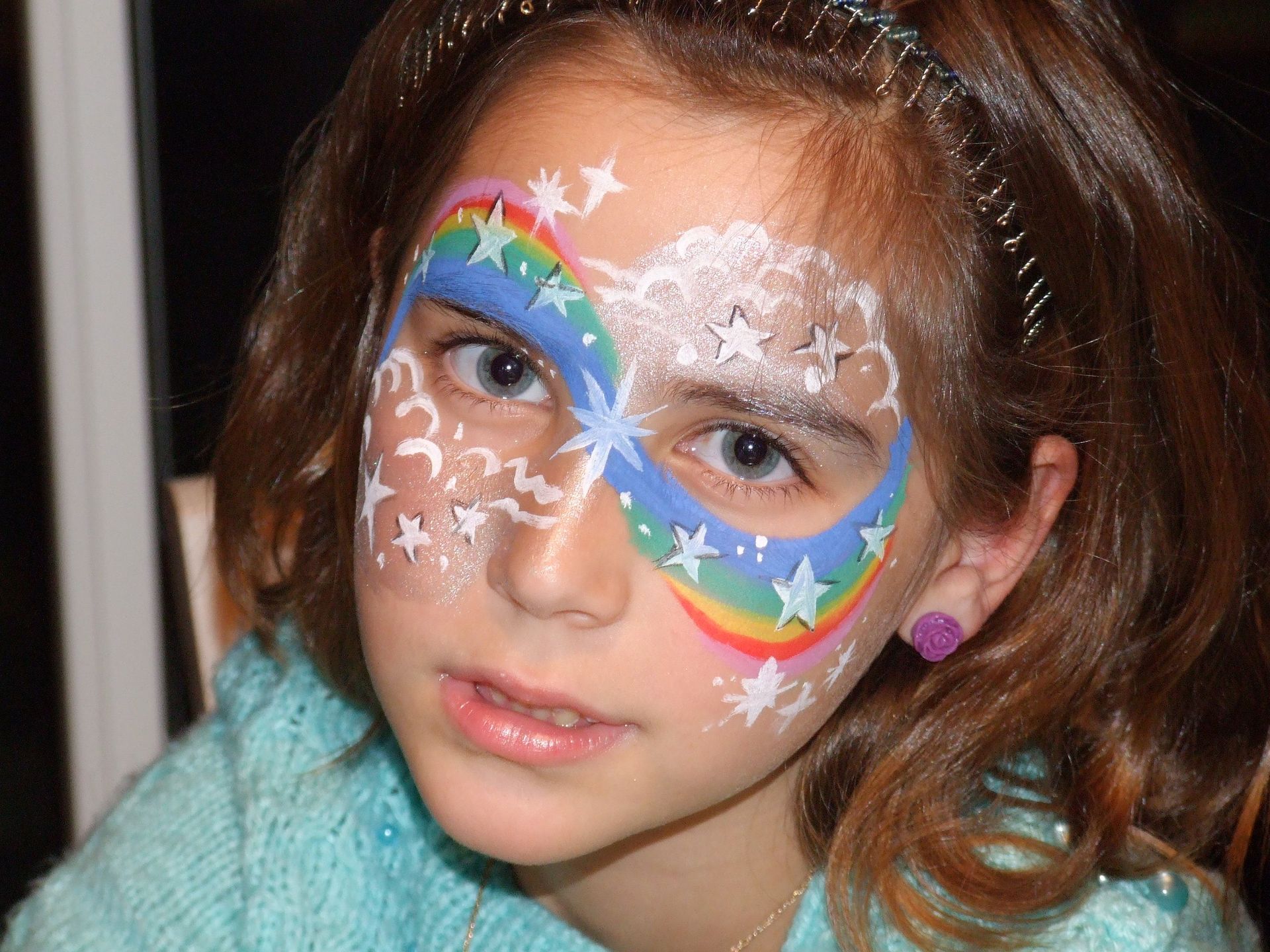 Enfant au visage maquillé de couleurs vives et de motifs d'étoiles blanches, portant un pull turquoise et regardant l'objectif.