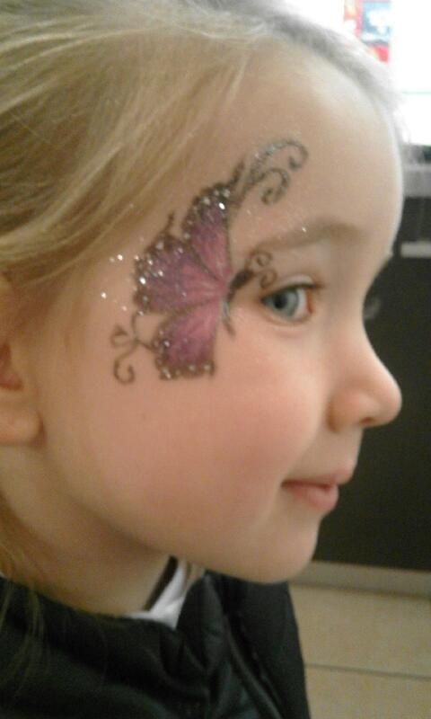 Enfant de profil avec un maquillage papillon violet pailleté sur la joue, à l'intérieur.