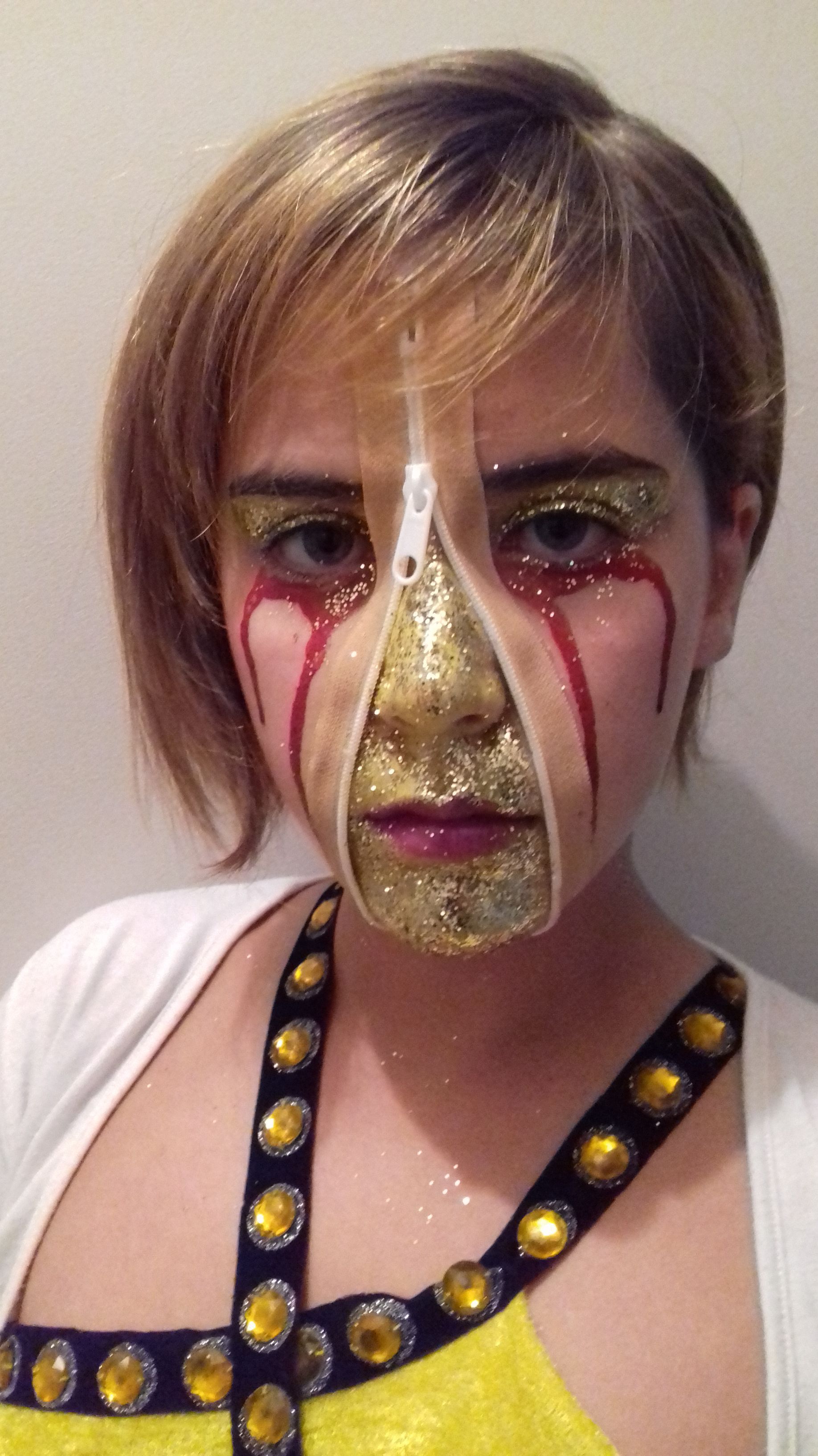 Personne au visage peint en doré avec des mèches rouges, vêtue d'un costume noir et jaune, regardant la caméra.