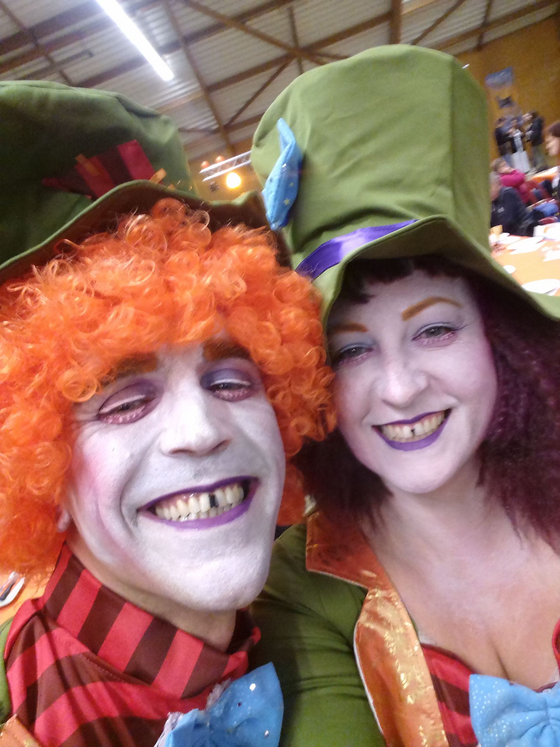 Deux artistes costumés et souriants, coiffés de chapeaux colorés et maquillés, lors d'un événement en intérieur
