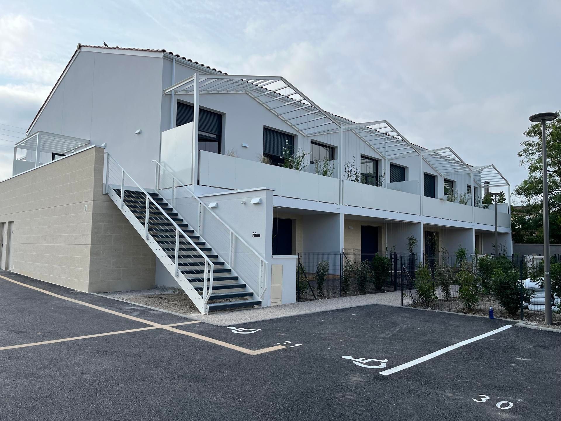 Un immeuble résidentiel moderne à logements multiples, de couleur blanche, avec un escalier extérieur en métal et des places de stationnement réservées aux personnes handicapées.
