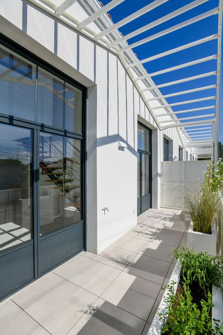 Bâtiment blanc avec un patio moderne, de grandes portes vitrées et une pergola à claire-voie sous un ciel bleu azur.
