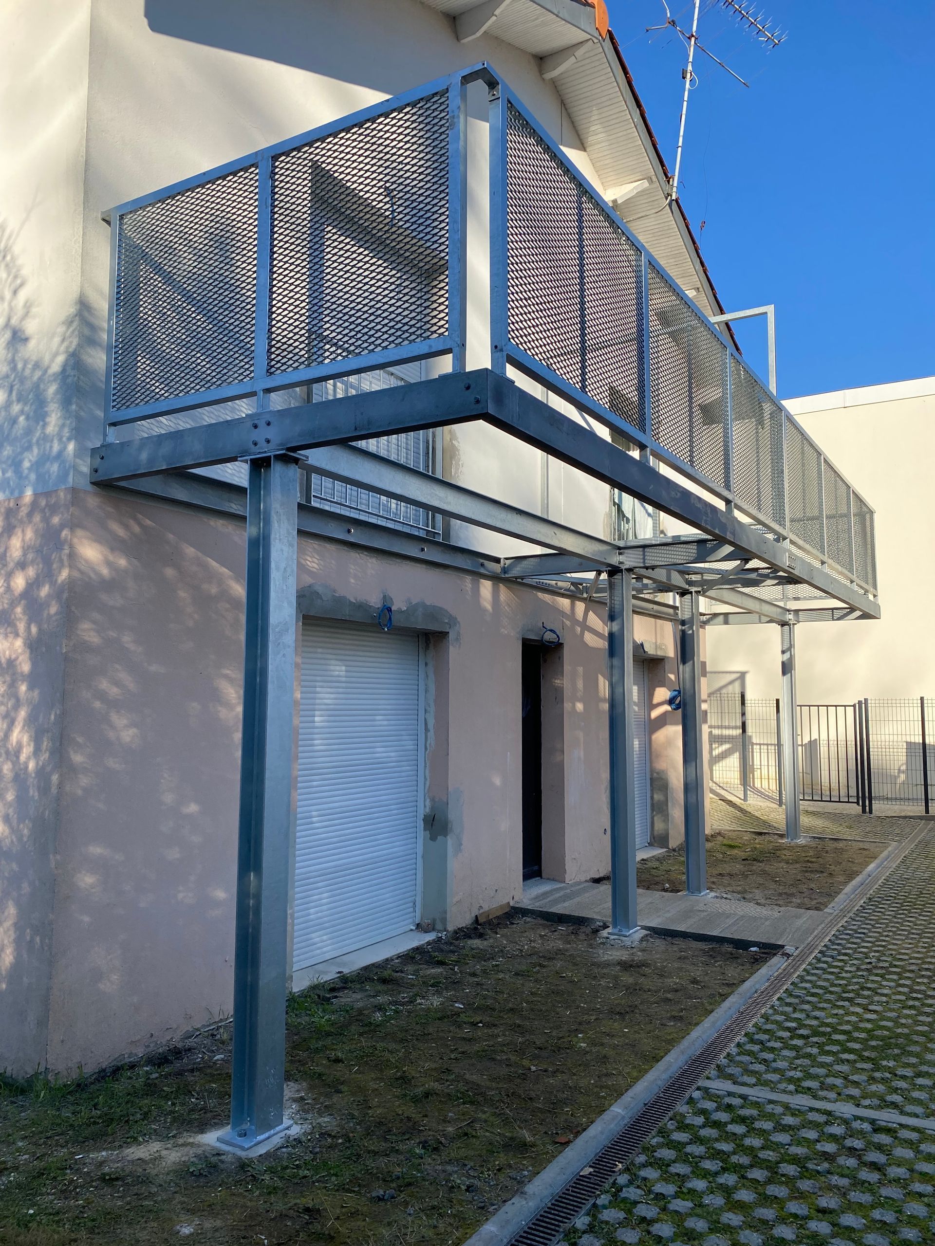 Un balcon surélevé en métal gris, doté d'une rambarde perforée, est soutenu par trois colonnes en acier adossées à la façade d'une maison.