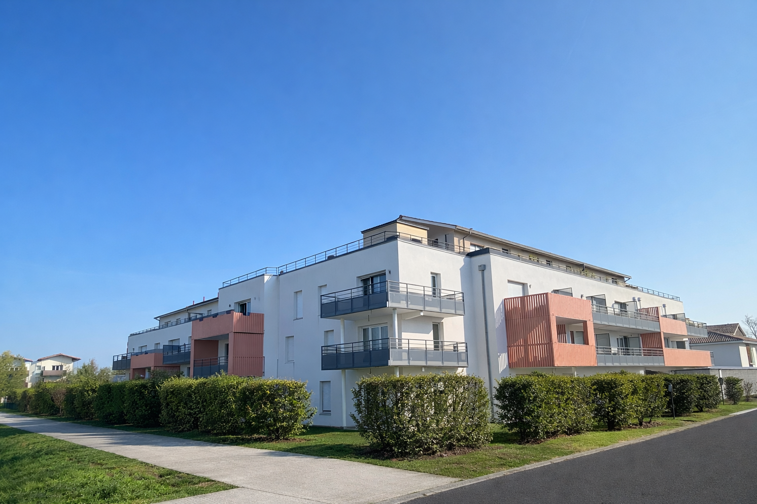 Un immeuble d'appartements blanc de trois étages, avec des balcons couleur pêche et des balustrades en métal foncé, se cache derrière une haie taillée.