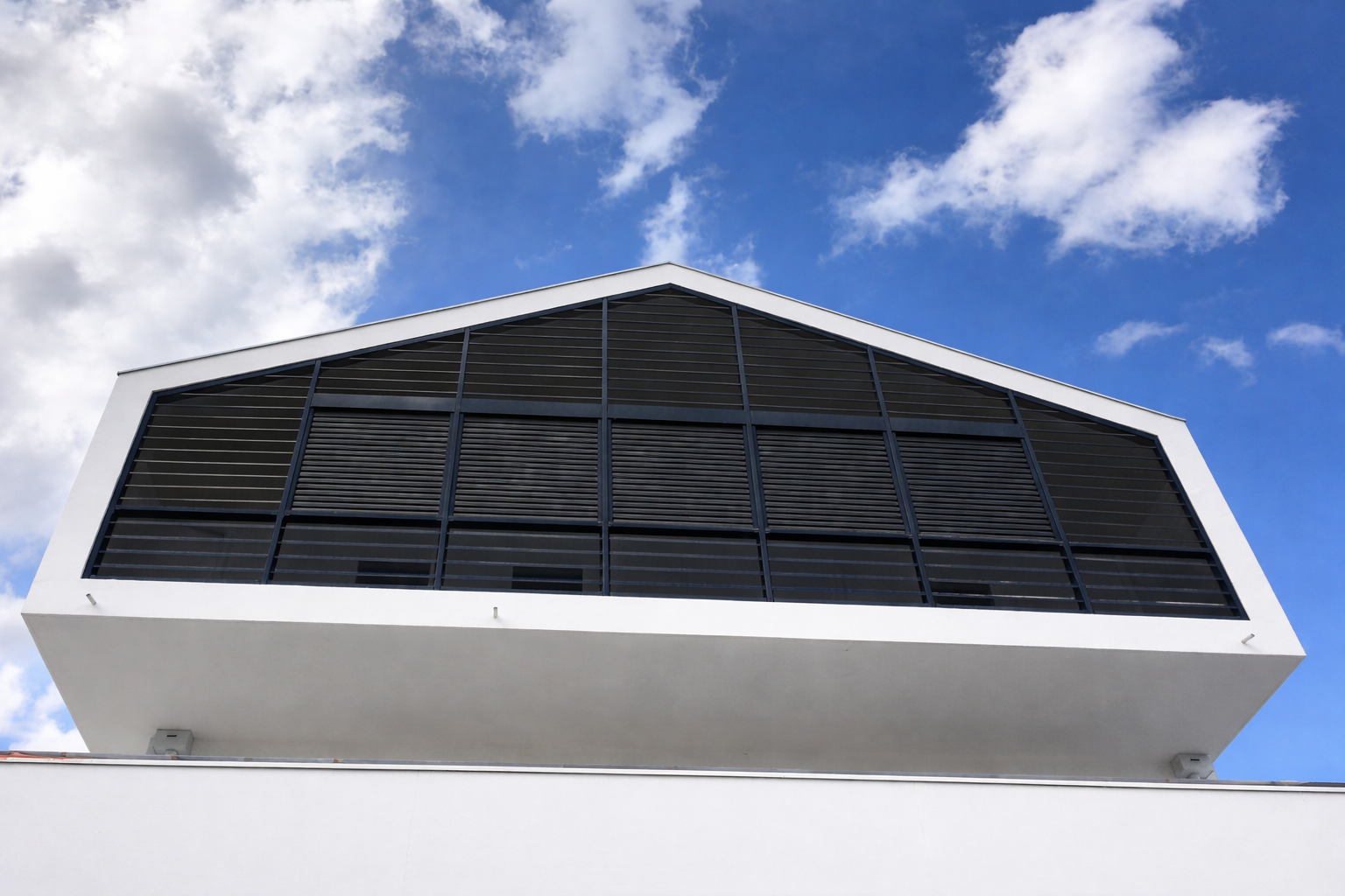 Une structure blanche à pignon, percée d'une grande fenêtre avec une grille noire, se détachant sur un ciel bleu vif et nuageux.
